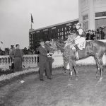 After running away with the Kentucky Derby at Churchill Downs, Assault, with jockey, Warren Mehrtens up, stands in the winner's circle wearing the traditional garland of roses. Trainer Max Hirsch is at left. Winner's share was $96,400.